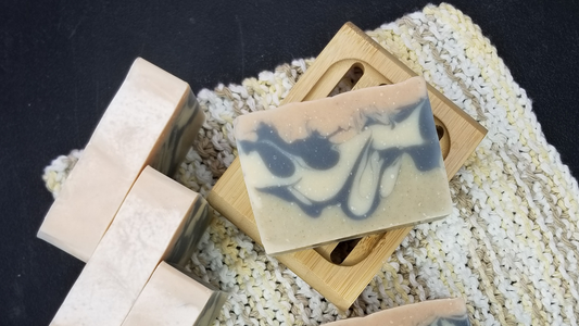 The top of several bars of artisan soap made with goat milk with a washcloth and the face of a bar made with charcoal, oatmeal, and nettle leaf powder sitting on a soap dish